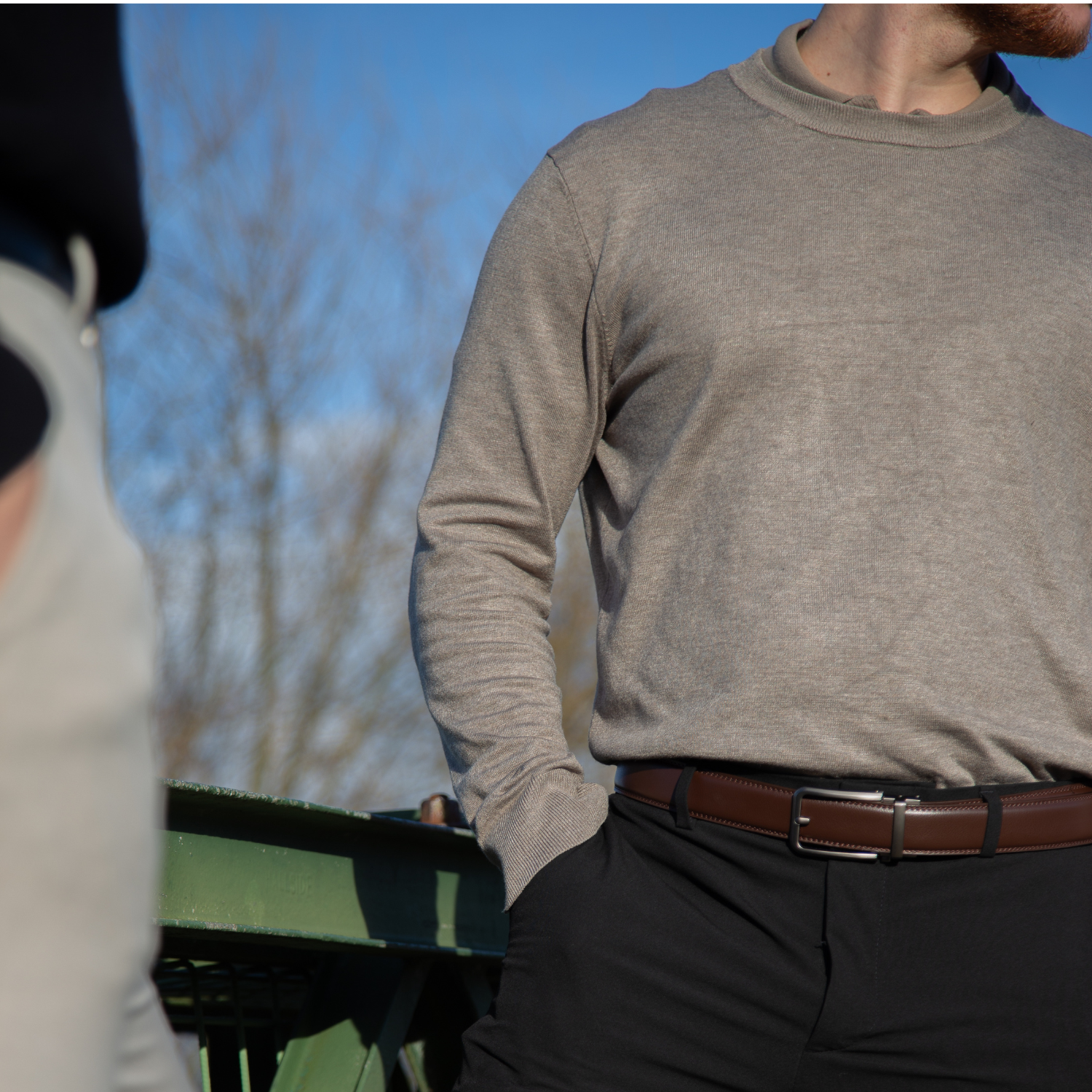 Een model draagt de bruine Buckle Up-riem terwijl hij op een brug staat, met een stijlvolle en tijdloze uitstraling.